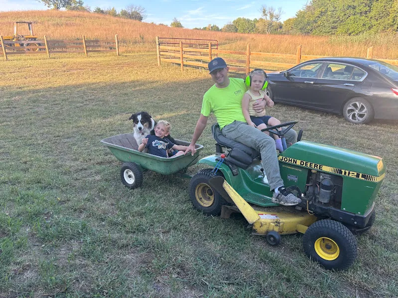 Man and two children riding a lawn tractor and trailer in a sunny yard.